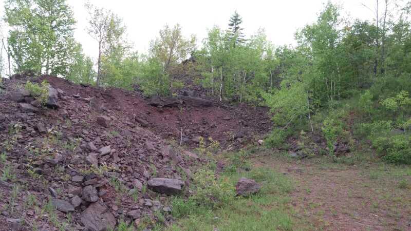 A rocky incline covered with scattered stones and patches of green vegetation, including small trees and shrubs, under a cloudy sky. Ispheming SBR trails mountain bike trail.