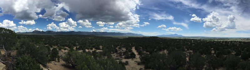 A panoramic view of a lush landscape featuring dense green vegetation and scattered trees, set against a backdrop of rolling mountains and a bright blue sky adorned with fluffy white clouds. The scene captures the natural beauty and tranquility of the outdoors. Phil's World mountain bike trail.