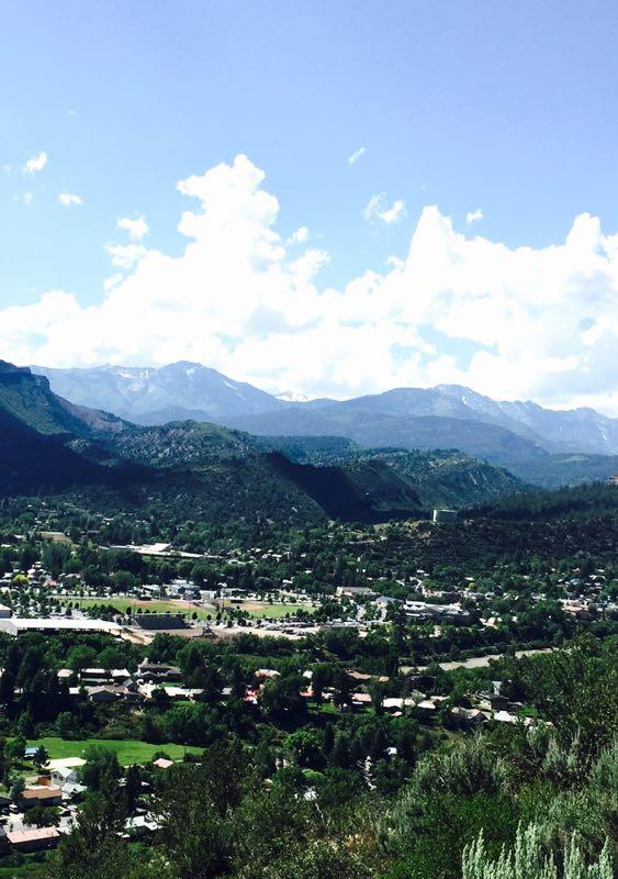 A panoramic view of a mountainous landscape under a blue sky with scattered clouds. In the foreground, a verdant valley is filled with trees and residential buildings, while expansive mountains rise in the background, some capped with snow. The scene captures the natural beauty and tranquility of the region. Telegraph Trail System mountain bike trail.