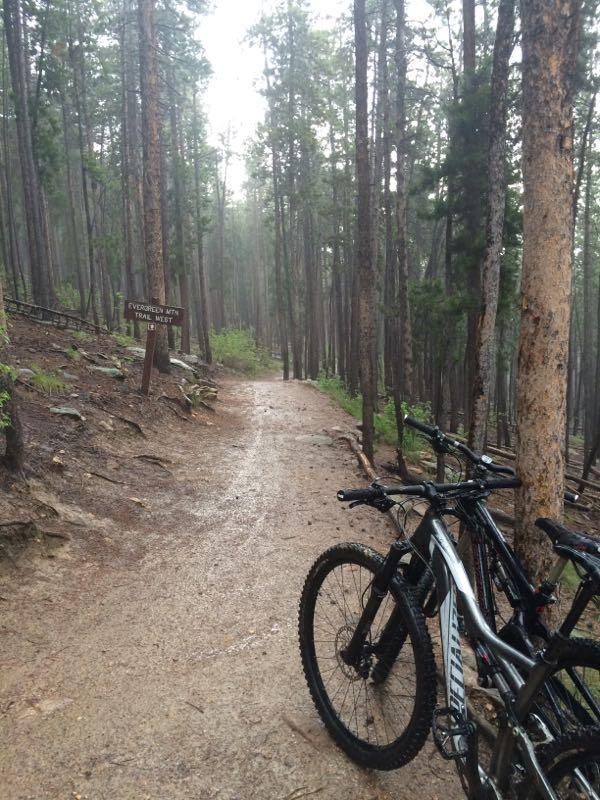 A misty forest trail in a mountainous area, with towering pine trees lining the path. Two mountain bikes are parked on the right side of the trail, and a wooden sign indicating the "Evergreen Mountain Trail" is visible nearby. The ground is damp, suggesting recent rain, and the atmosphere is calm and serene. 3 Sisters / Alderfer mountain bike trail.