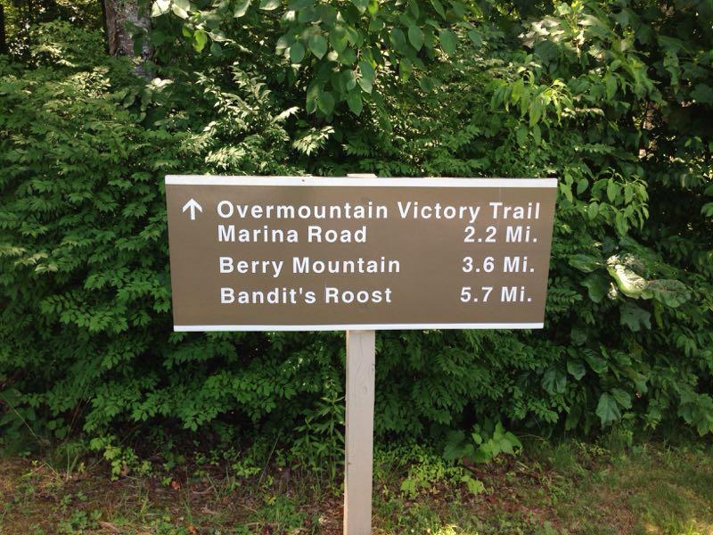 A directional sign indicating distances to the Overmountain Victory Trail (2.2 miles), Berry Mountain (3.6 miles), and Bandit's Roost (5.7 miles), surrounded by greenery. Kerr Scott Trails mountain bike trail.