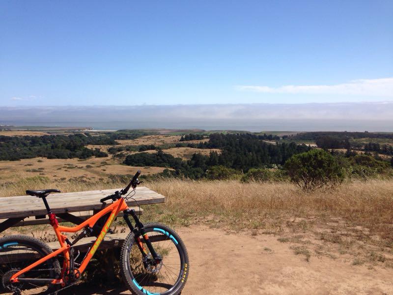 A vibrant orange mountain bike is leaning against a rustic wooden picnic table, overlooking a scenic landscape of rolling hills, greenery, and a distant view of a fog-covered coastline under a clear blue sky. Wilder Ranch State Park mountain bike trail.
