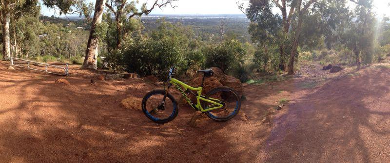 A bright green mountain bike is parked on a dirt trail surrounded by trees and shrubs, with a scenic view of the landscape in the background. The terrain is rugged, featuring rocky outcrops and a mix of natural flora. Kalamunda Railway Heritage Trail mountain bike trail.