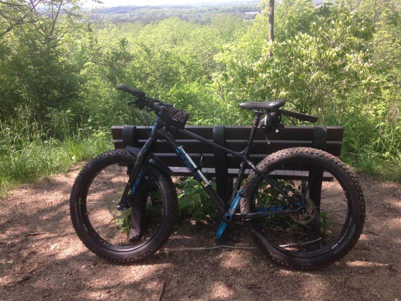 Mountain bike parked beside a wooden bench, overlooking a lush green landscape with trees and hills in the distance on a sunny day. Levis Mounds mountain bike trail.