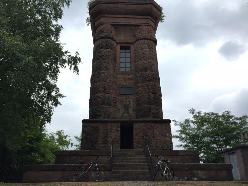 A tall stone tower with a staircase leading to the entrance, surrounded by trees and under a cloudy sky. Two bicycles are parked at the base of the tower. Tower Trail mountain bike trail.