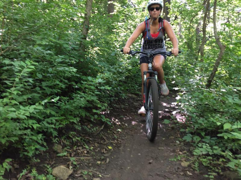 A person riding a mountain bike on a narrow dirt trail surrounded by dense greenery. The individual is wearing a helmet and sunglasses, and their clothing reflects an active outdoor vibe. The setting suggests a sunny day, highlighting the natural environment. Shawnee Mission Park mountain bike trail.