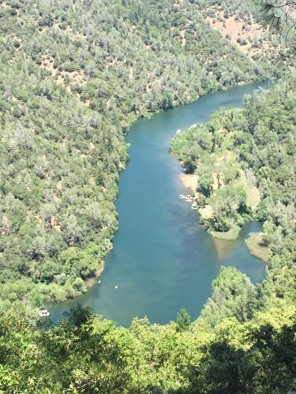 Aerial view of a winding river surrounded by lush greenery, with patches of land and trees on either side. Small boats are visible on the water, showcasing a tranquil and picturesque natural landscape. Foresthill Divide mountain bike trail.