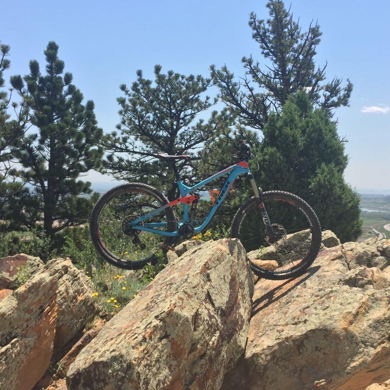 A colorful mountain bike is positioned on a rocky outcrop, surrounded by green trees and wildflowers, under a clear blue sky. The landscape showcases a scenic view of hills and valleys in the background. Red Rocks / Dakota Ridge mountain bike trail.