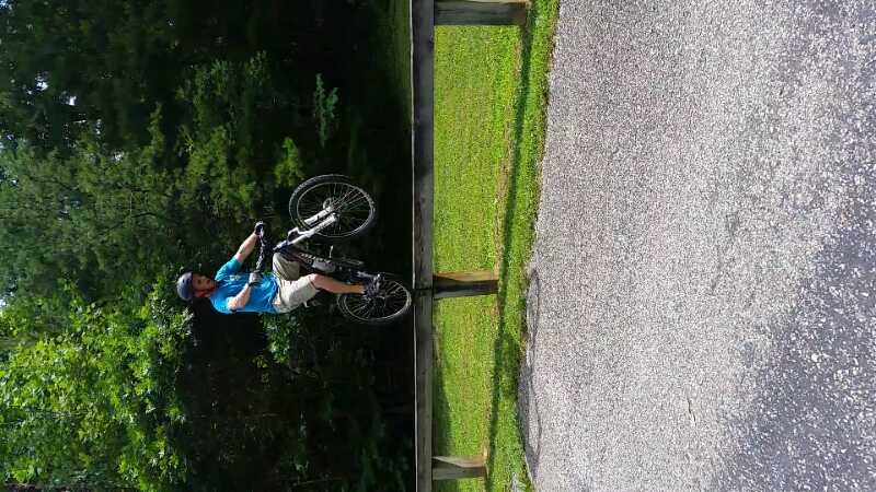A person performing a bicycle stunt, riding on the front wheel, next to a wooden railing. The scene is set in a grassy area bordered by trees, with a gravel path alongside. The individual is wearing a blue shirt and a helmet. Tsali Recreation Area mountain bike trail.