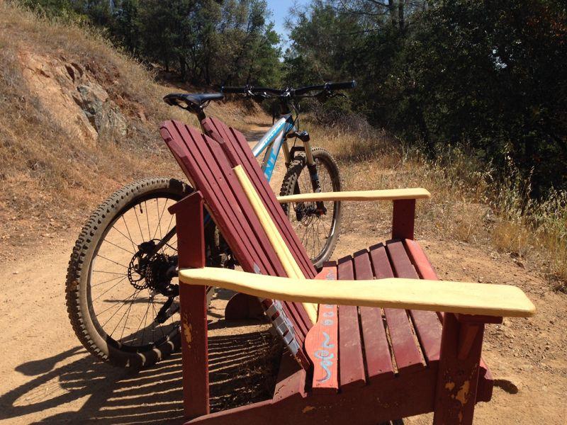 A mountain bike resting against a colorful wooden bench along a dirt trail, surrounded by trees and dry grass. The sunny outdoor setting suggests a peaceful spot for resting after a ride. Stagecoach / Flood / Manzanita mountain bike trail.