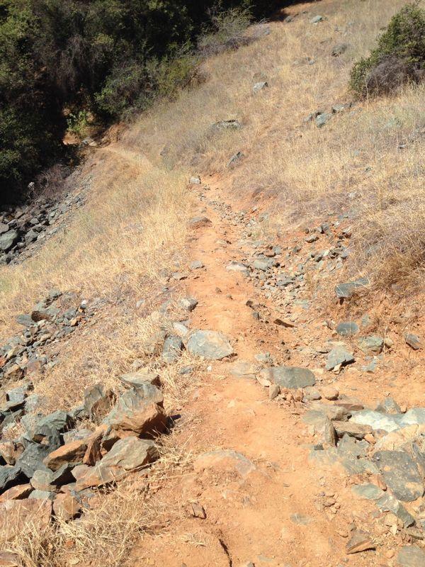 A winding dirt path through dry, rocky terrain, flanked by sparse vegetation and dry grass. The path descends along a hillside, with scattered stones and patches of earth visible. Stagecoach / Flood / Manzanita mountain bike trail.