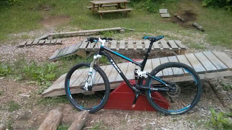 A mountain bike rests on a wooden obstacle course set in a grassy area, surrounded by trees. The bike features a black frame with blue accents and is positioned near various wooden ramps and platforms. In the background, a picnic table is visible, adding to the outdoor setting. Hardwood Ski and Bike mountain bike trail.