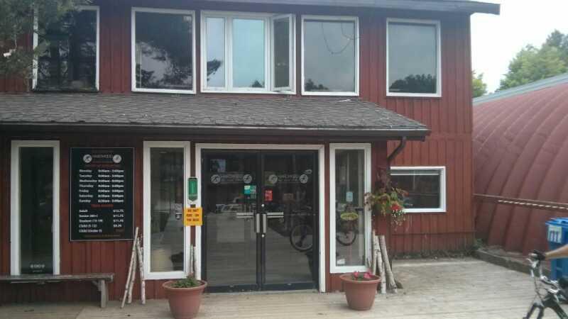Front view of a two-story red wooden building with large windows and a double-door entrance. A signboard on the left displays operational hours and services. Potted plants adorn the entrance, and there are wooden benches and a bike nearby, indicating a welcoming atmosphere. Hardwood Ski and Bike mountain bike trail.