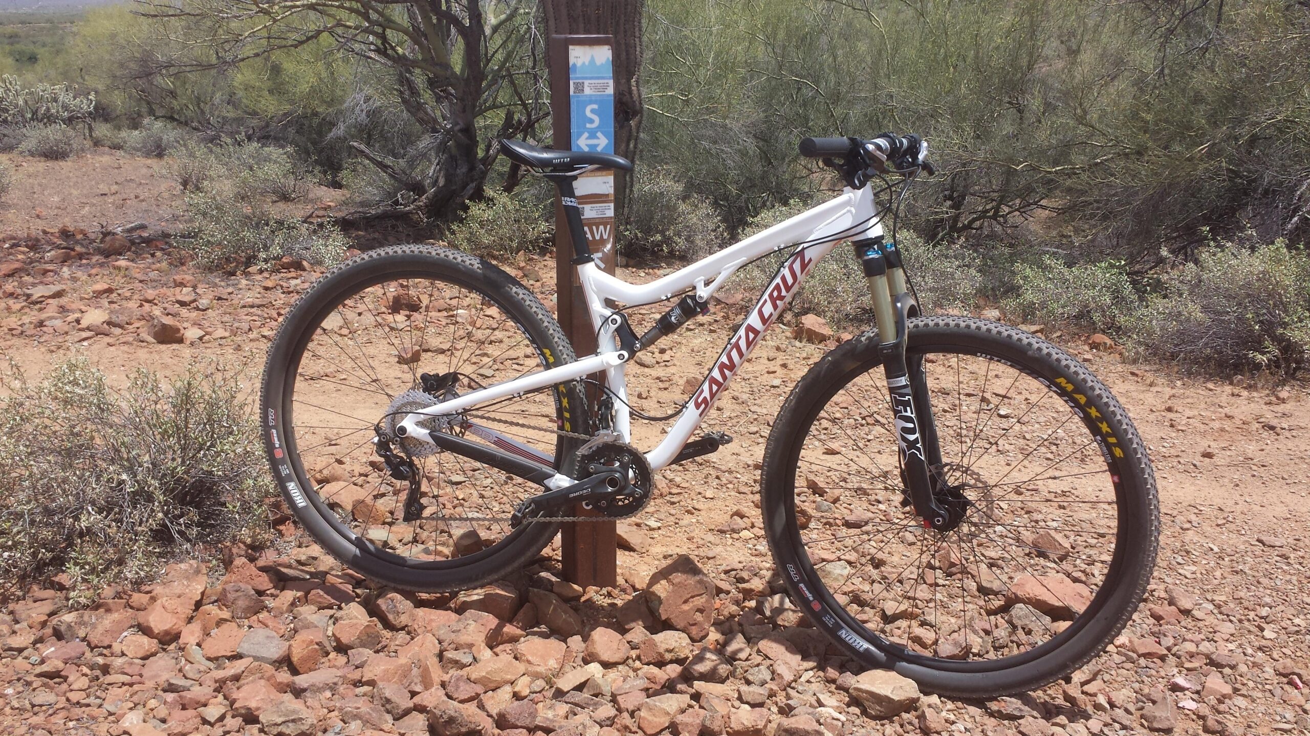 Santa Cruz Tallboy: A white mountain bike leaning against a trail sign amidst a rocky, desert landscape with sparse vegetation. The sign indicates a mountain biking route, and the sun casts a bright light over the scene.