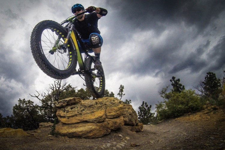 A cyclist performing a jump on a mountain bike over a rocky ledge, with a dramatic sky in the background. Trees and shrubs flank the trail, indicating a rugged outdoor environment.