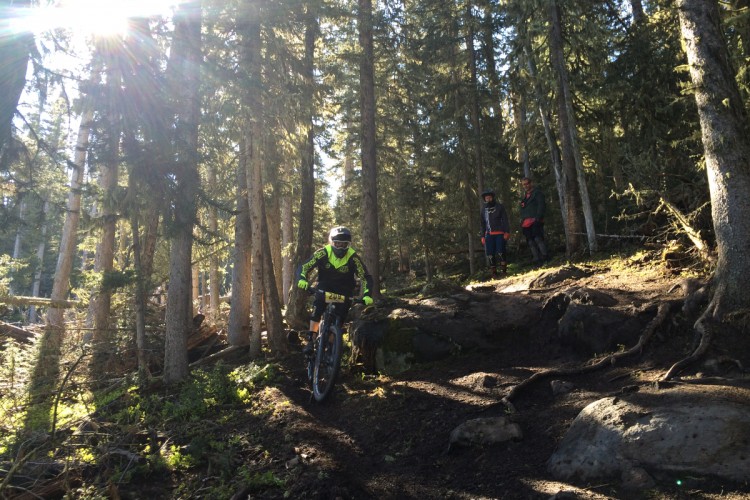 A mountain biker rides along a rocky trail in a forest, surrounded by tall trees and dappled sunlight. Two individuals stand on the trail to the right, observing the biker as they navigate the uneven terrain.