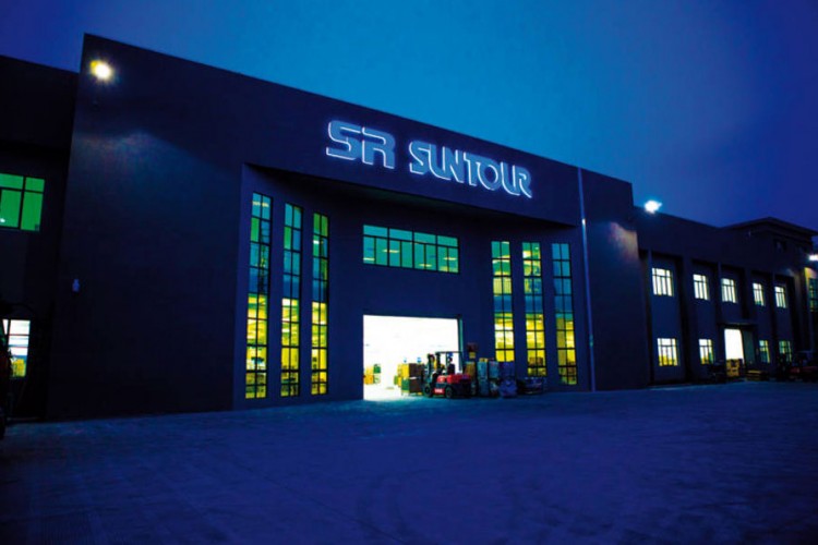 An industrial building at dusk with the illuminated sign "SR SUNTOUR" prominently displayed. The structure features large windows that are glowing with yellow light, suggesting activity inside, while forklifts and stacks of materials are visible near the entrance.