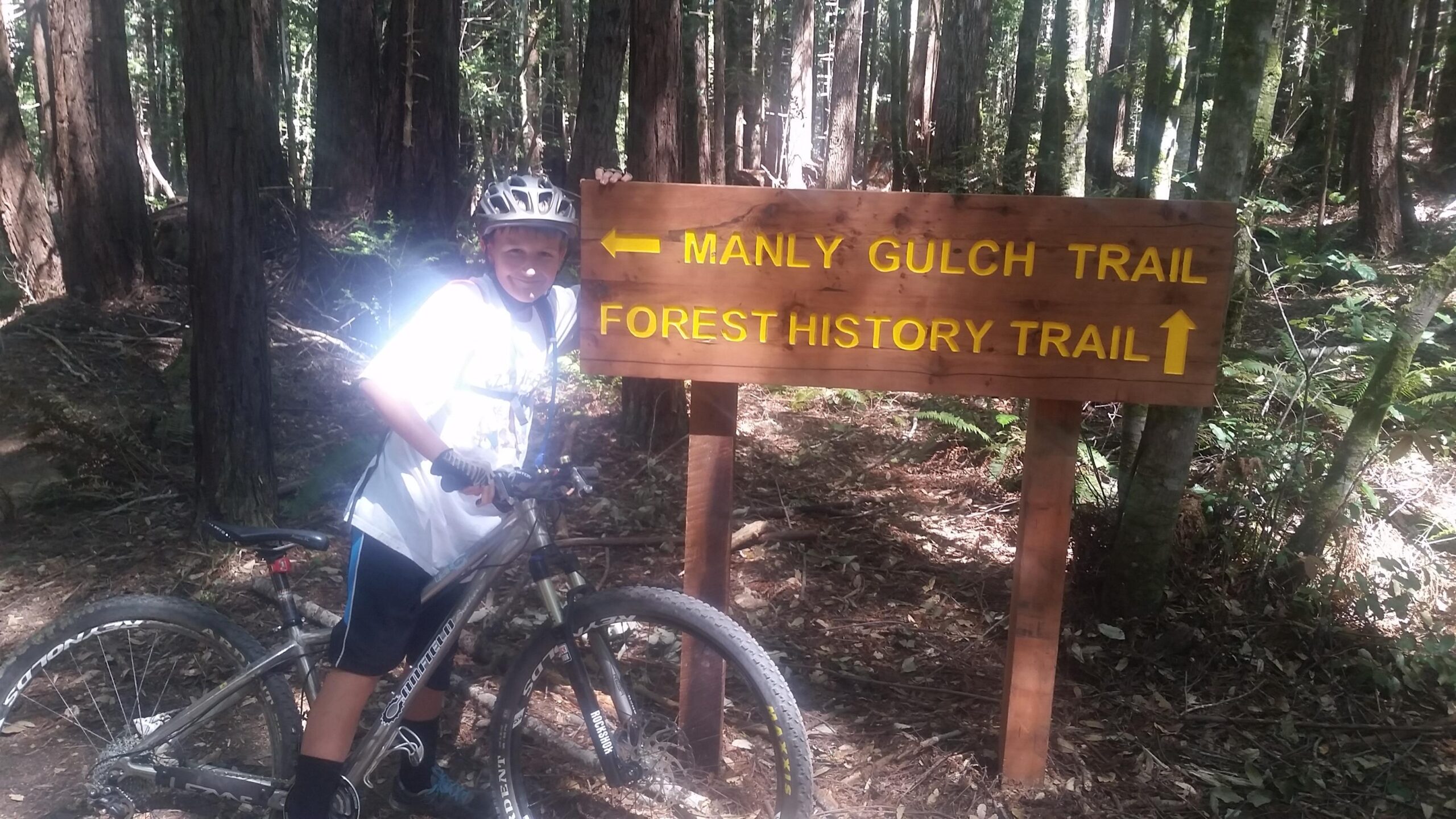 A young boy with a bicycle stands next to a trail sign in a forest, indicating directions to Manly Gulch Trail and Forest History Trail. Tall trees and lush greenery surround the area. Manly Gulch mountain bike trail.