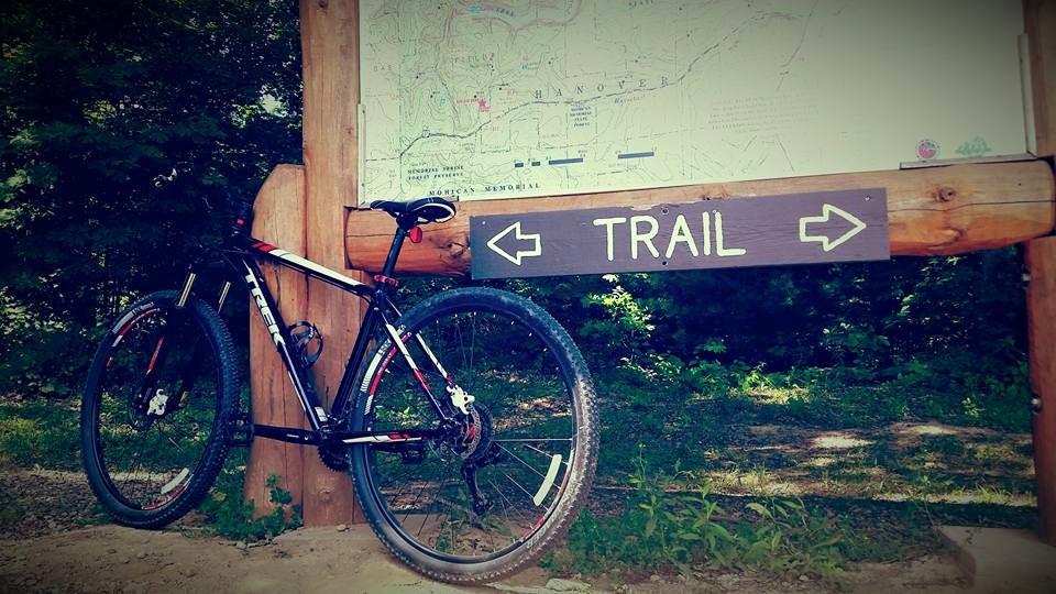 A mountain bike resting against a wooden trail sign, with arrows indicating directions, and a map of the surrounding area visible above the sign. The setting is surrounded by green foliage, suggesting a natural outdoor environment. Mohican mountain bike trail.