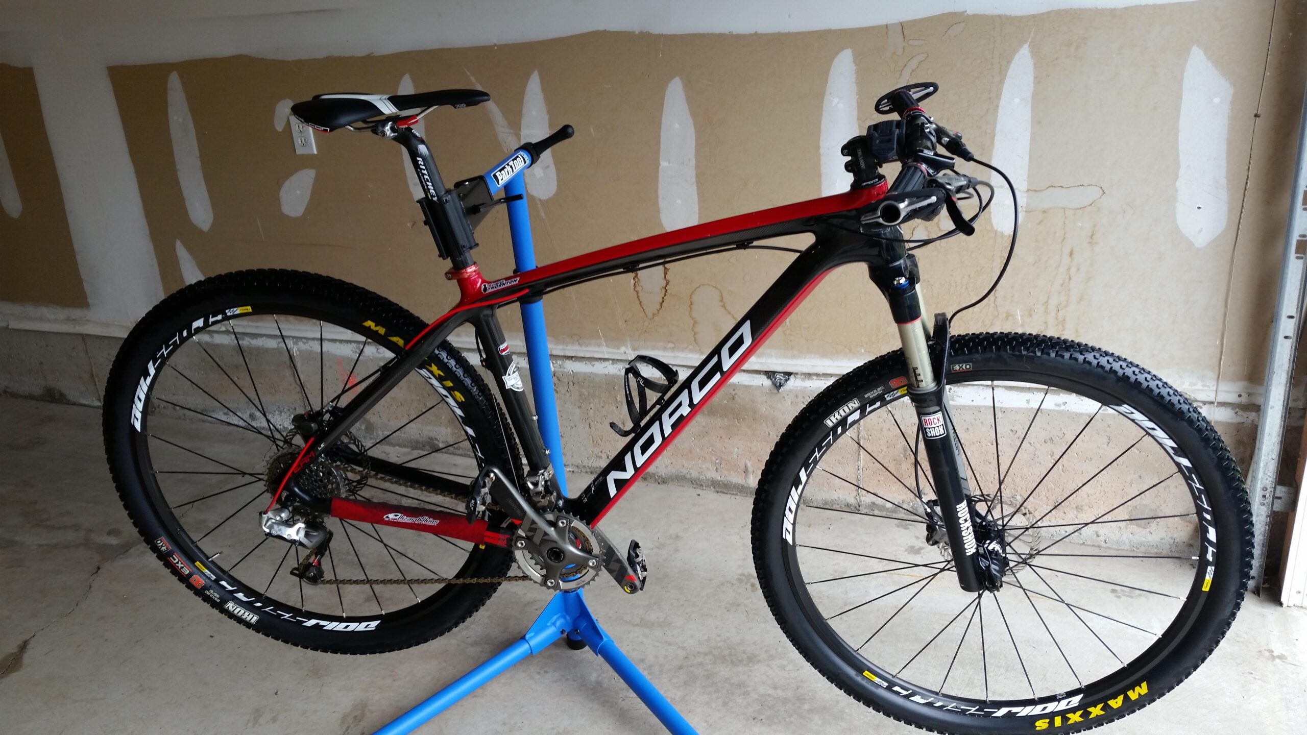 Norco Team 9 LE: A red and black Norco mountain bike mounted on a blue repair stand, with its front wheel showing Maxxis branding on the tire. The bike is positioned in a garage with unfinished walls in the background.
