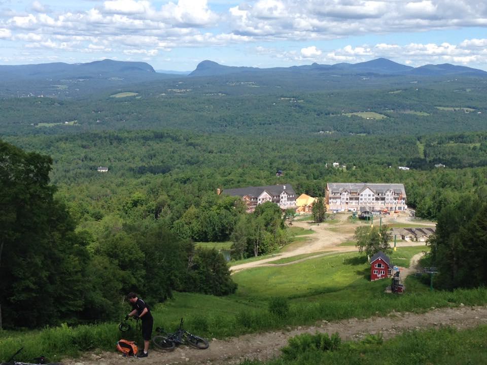 A scenic view from a mountain summit overlooking a lush green valley, with distant mountains in the background. In the foreground, a person is adjusting their bicycle gear next to a tan backpack, while several buildings and a ski lift are visible in the lower landscape. The sky is partly cloudy, adding to the vibrant atmosphere of the outdoor setting. Kingdom Trails mountain bike trail.