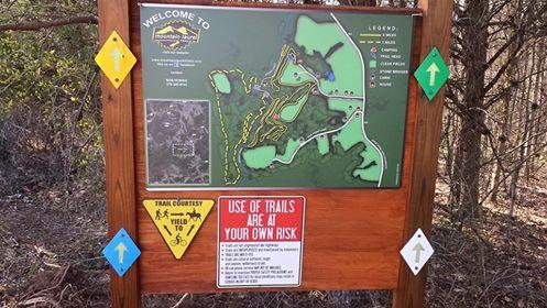 A wooden sign displaying a map of Mountain Park, with trail information and safety warnings. The map outlines various trails and their ratings, and includes symbols indicating bike and trail user instructions. Text warns that trail use is at one's own risk. Surrounding trees and nature are visible in the background. Mountain Laurel Trails mountain bike trail.