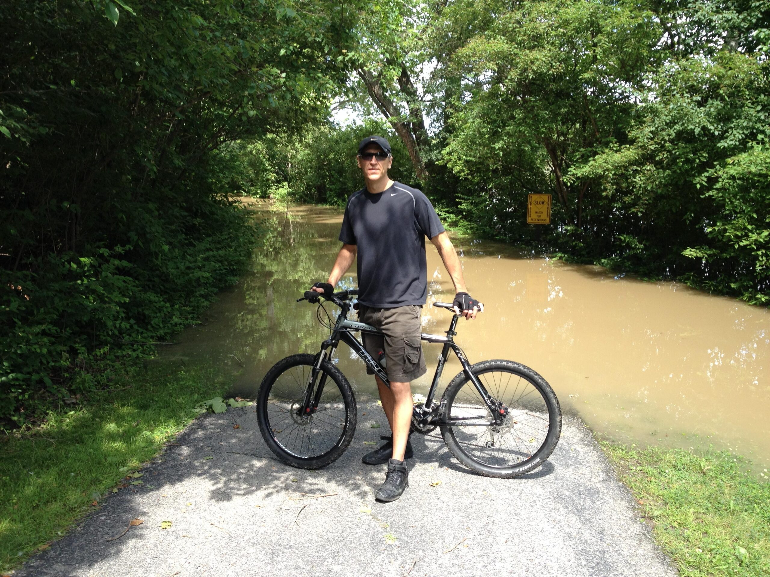 Trek 6000: A person stands next to a black mountain bike on a path that is partially submerged in muddy water, surrounded by dense greenery. A "SLOW" sign is visible in the background, indicating caution due to the flooding. The scene suggests a recent rise in water levels along the trail.