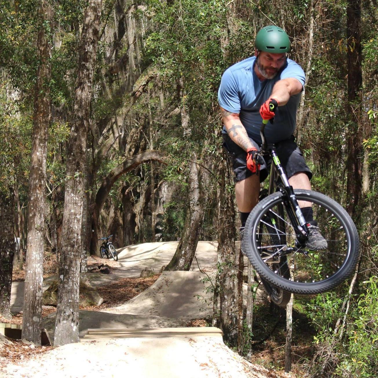 A mountain biker performing a jump on a dirt trail in a wooded area. The rider, wearing a green helmet and gloves, is airborne above a dirt ramp, surrounded by trees and a winding bike path. Another bicycle can be seen in the background. Santos mountain bike trail.