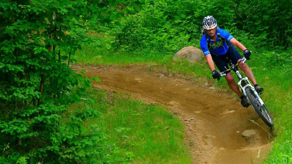 A mountain biker navigating a curved trail through lush green foliage, wearing a blue jersey and helmet. The cyclist is leaning into the turn, demonstrating skill and agility on the bike. Kingdom Trails mountain bike trail.