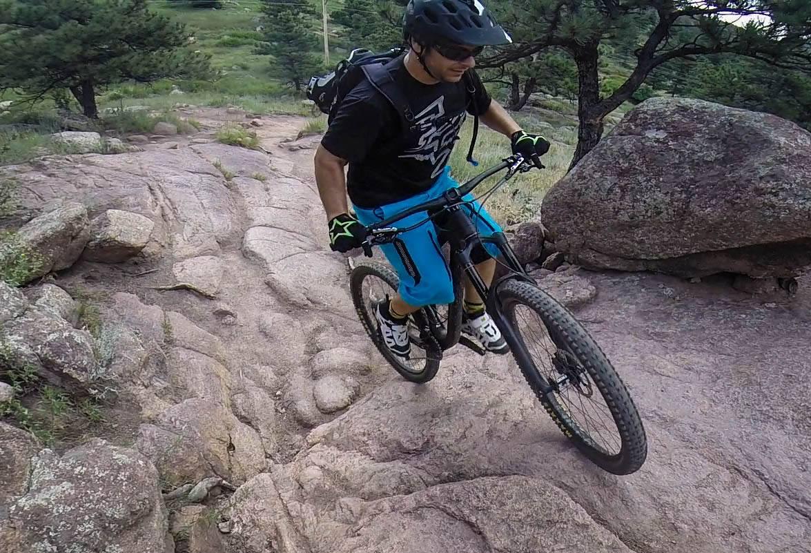 Santa Cruz Nomad Carbon 27.5: A mountain biker navigating a rocky trail, wearing a helmet and protective gear. The rider is dressed in a black shirt and blue shorts, focusing on maneuvering a mountain bike over uneven terrain with trees and shrubbery in the background.