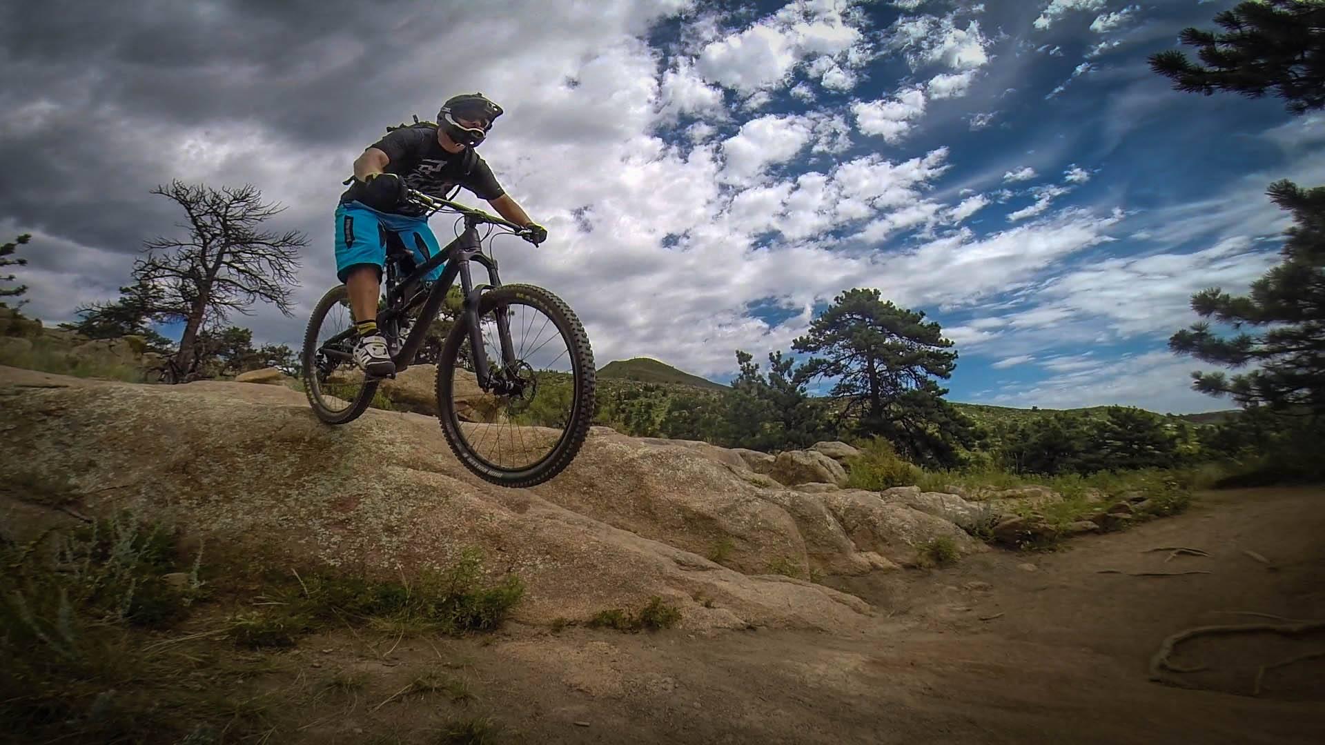 Santa Cruz Nomad Carbon 27.5: A mountain biker in a black t-shirt and blue shorts is performing a jump over a rocky terrain. The background features trees and a sky with scattered clouds, suggesting an outdoor trail.