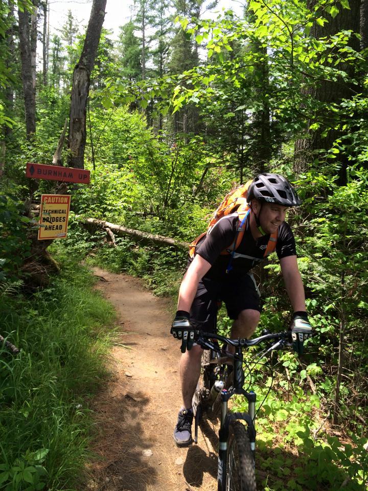 A mountain biker riding on a narrow trail through a lush green forest. Signs in the background indicate the trail name "Burnham Up" and caution about technical bridges ahead. Sunlight filters through the trees, adding warmth to the scene. The rider is wearing a helmet, gloves, and a backpack, focused on navigating the path. Kingdom Trails mountain bike trail.