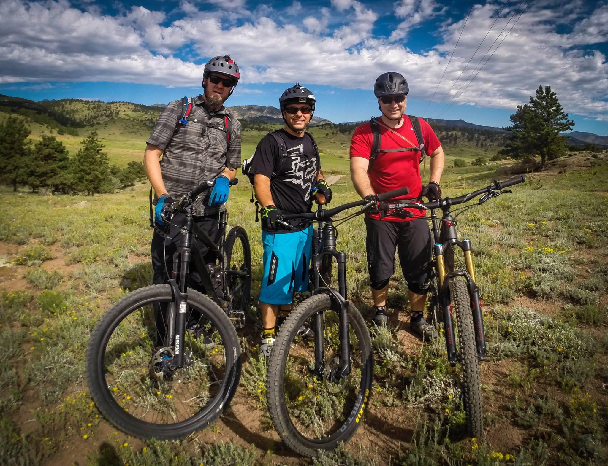 Santa Cruz Nomad Carbon 27.5: Three mountain bikers pose outdoors, each with a mountain bike, against a scenic backdrop of green hills and a blue sky with scattered clouds. The first rider wears a plaid shirt and black gloves, the second is in a black shirt and blue shorts, and the third is in a red shirt. They stand together in a grassy area with some wildflowers.