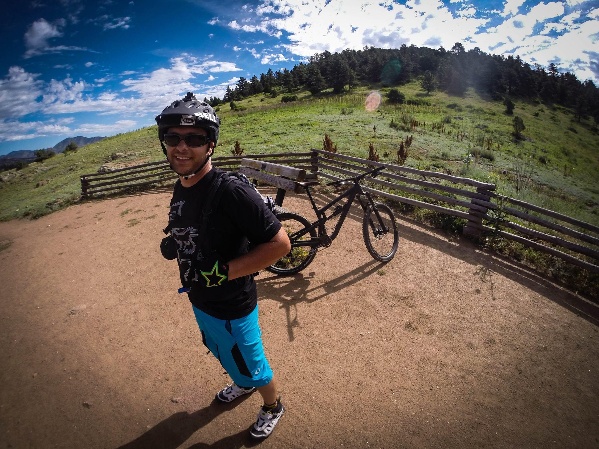 Santa Cruz Nomad Carbon 27.5: A mountain biker stands on a dirt path, wearing a helmet and sunglasses, with a black mountain bike beside him. He is smiling and dressed in a black t-shirt and blue shorts. The background features a green, hilly landscape under a mostly blue sky with scattered clouds and a wooden fence.