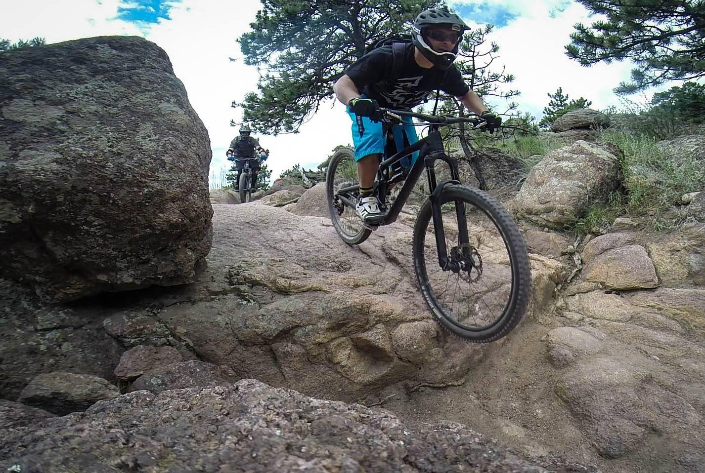 Santa Cruz Nomad Carbon 27.5: Two mountain bikers navigate a rocky trail surrounded by trees under a cloudy sky. The rider in the foreground is airborne as they jump over a large rock, demonstrating skill and agility on their bike, while the second rider follows closely behind.