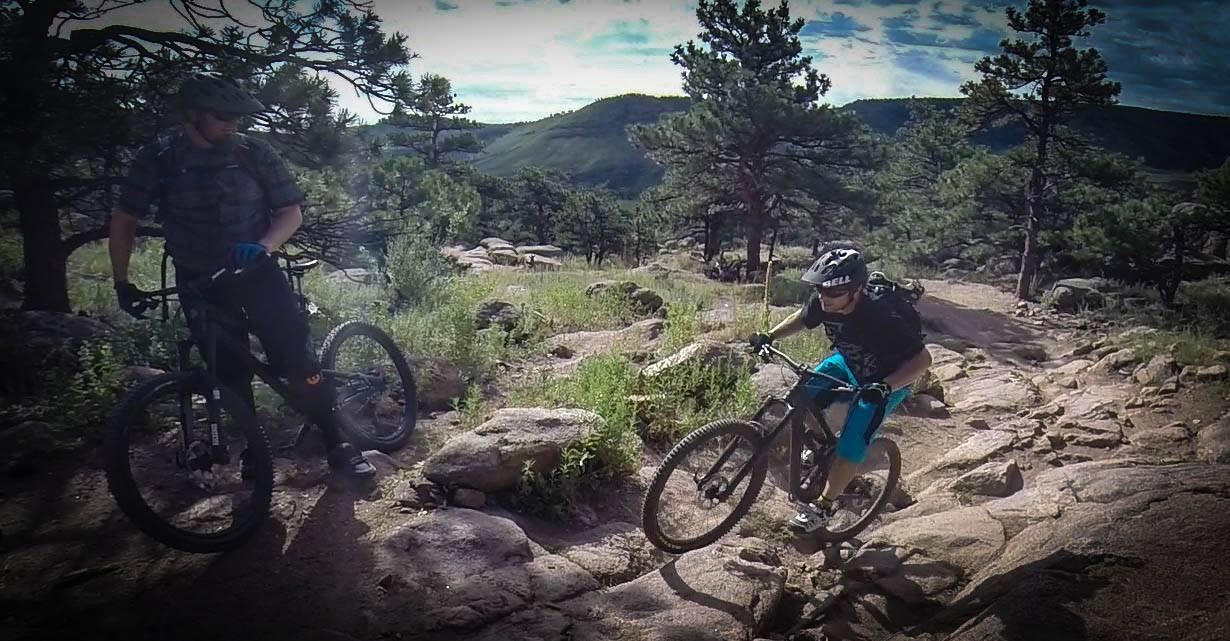 Santa Cruz Nomad Carbon 27.5: A child navigating a rocky mountain biking trail, wearing a helmet and riding a bicycle, while an adult stands nearby on their bike, surrounded by trees and scenic mountainous terrain.