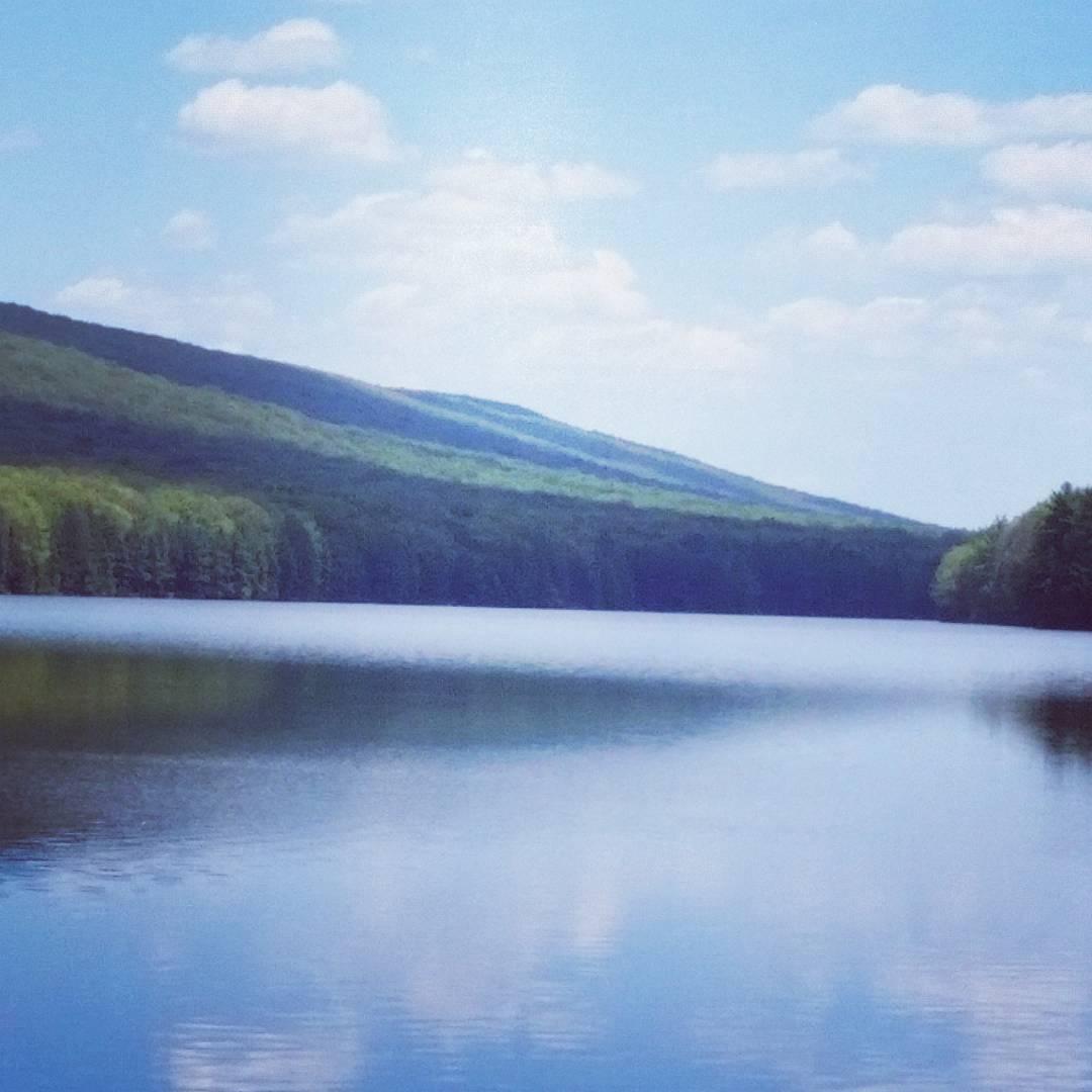 A serene lake surrounded by lush greenery and rolling hills under a clear blue sky with fluffy white clouds. The calm water reflects the landscape, creating a picturesque and tranquil scene. Roaring Creek Watershed mountain bike trail.