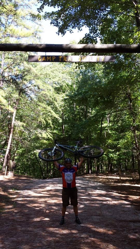 A cyclist in a helmet stands on a dirt path, joyfully raising a mountain bike above their head. In the background, a wooden sign reads "Camp Tranquility," framed by lush green trees and sunlight filtering through the leaves. Coldwater Mountain mountain bike trail.