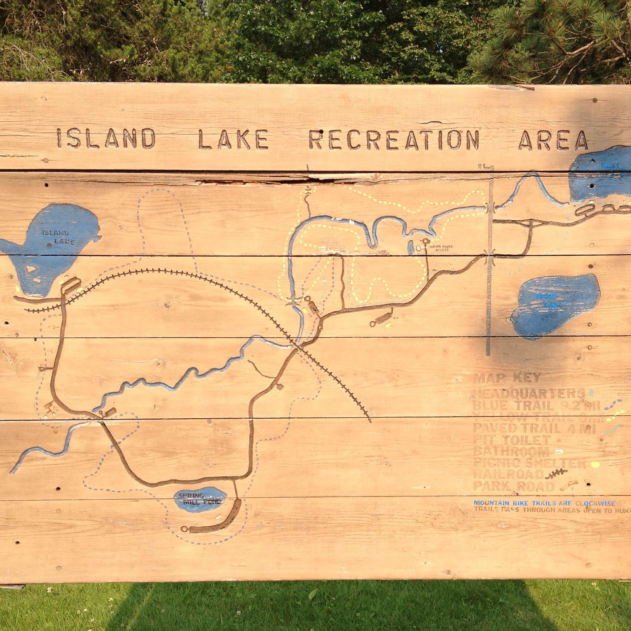 A wooden sign depicting a map of the Island Lake Recreation Area, featuring labeled lakes, trails, and facilities. The map includes key features such as headquarters, restrooms, and picnic areas, with marked blue trails for biking and walking. The background shows greenery, indicating the natural setting of the park. Island Lake mountain bike trail.