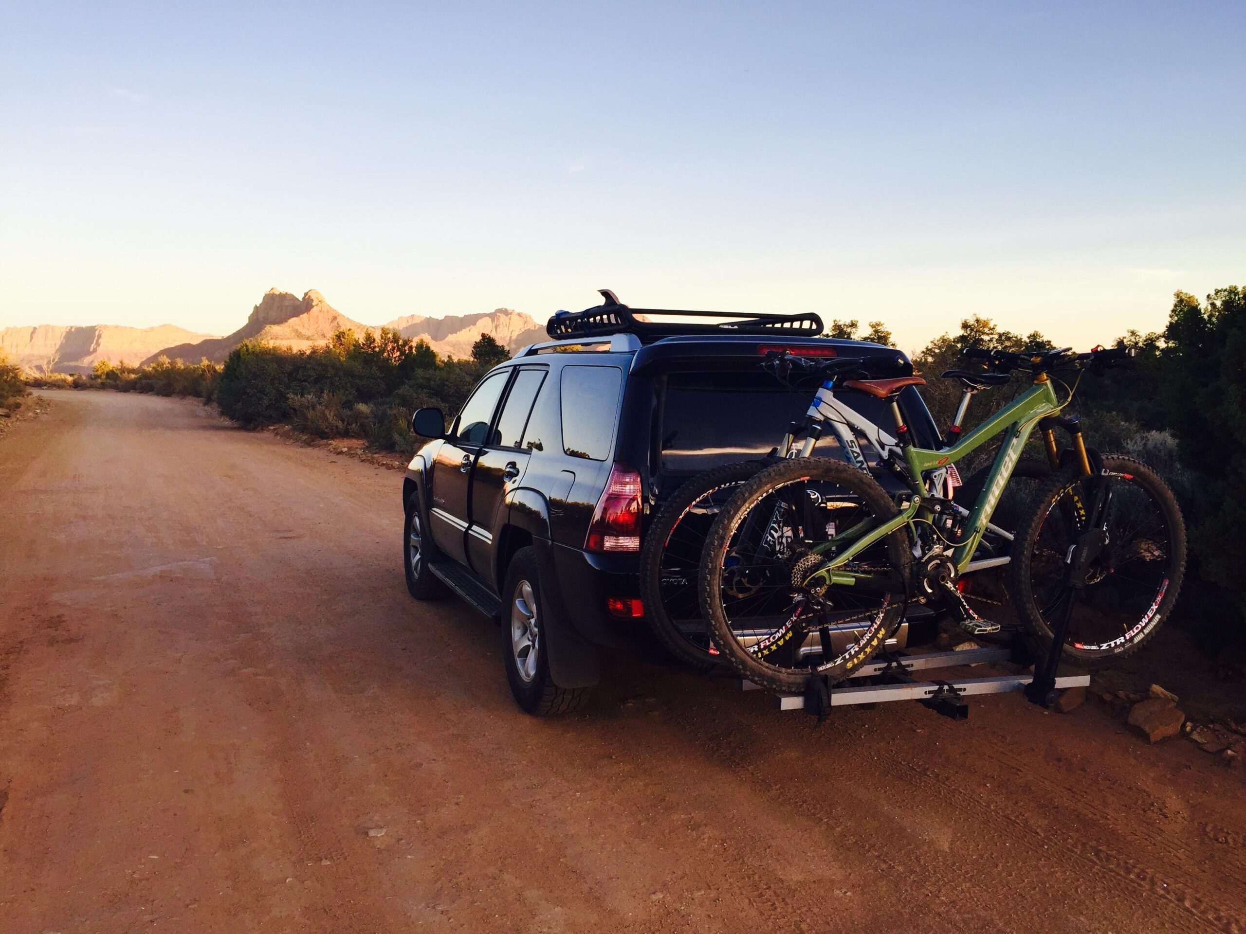 Santa Cruz Bronson: An SUV parked on a dirt road, with two mountain bikes secured on a bike rack attached to its rear. The background features rugged mountains and a clear sky during sunset. The scene conveys an adventurous outdoor setting.
