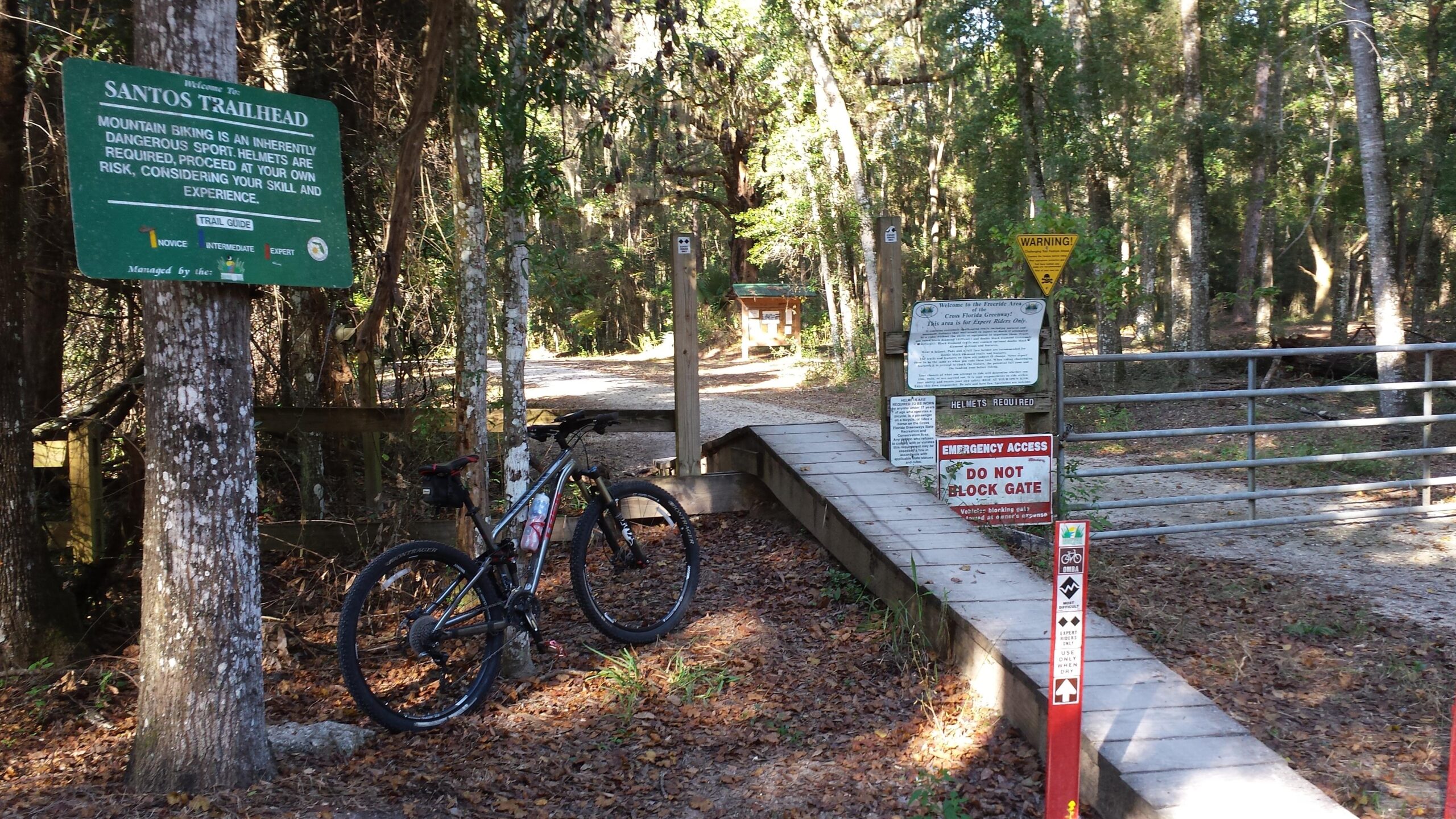 A mountain bike is parked next to the Santos Trailhead entrance, featuring multiple signs that provide information about mountain biking safety, helmet requirements, and trail skill levels. The setting is surrounded by trees, with a gravel path leading further into the woods. Santos mountain bike trail.