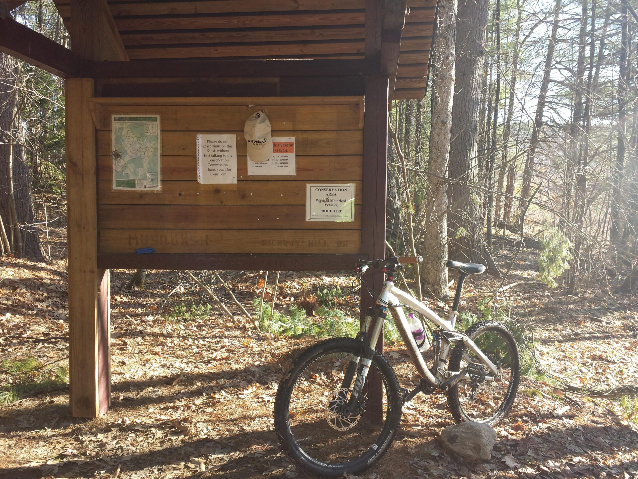 A mountain bike rests against a wooden informational sign in a wooded area, surrounded by fallen leaves and trees. The sign features a map, various notices and rules, while a sunlit path leads into the forest in the background. Musquash Conservation Area mountain bike trail.