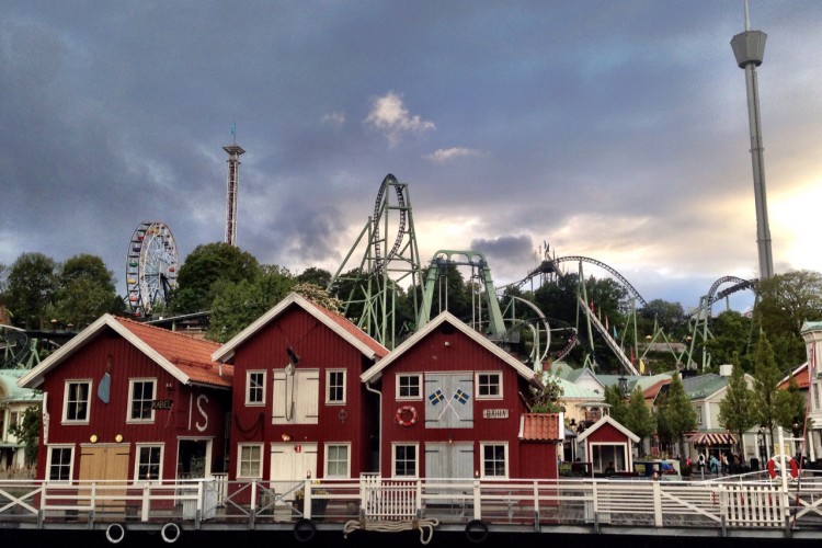 Image of a waterfront scene featuring charming red wooden houses with white trim. In the background, amusement park rides, including roller coasters and a Ferris wheel, are visible against a moody sky. The reflection of the buildings can be seen in the water, creating a picturesque setting.