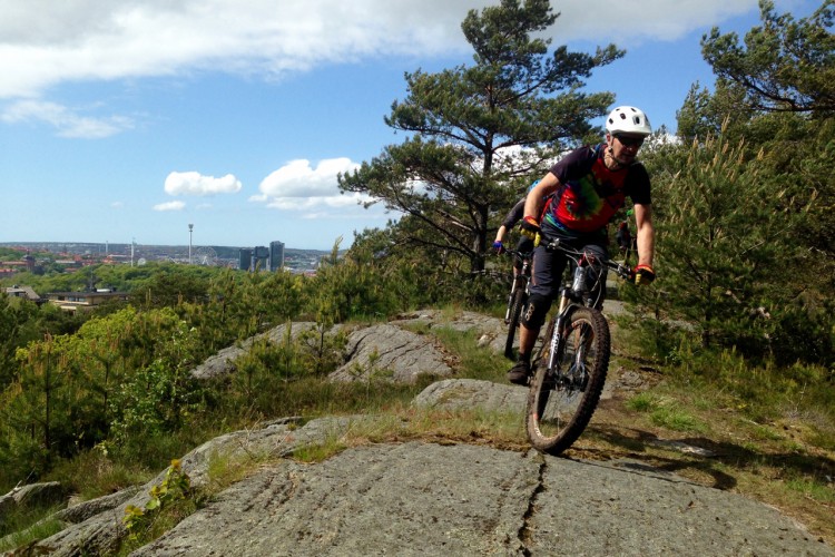 A mountain biker rides along rocky terrain with trees in the background, overlooking a cityscape with buildings and a Ferris wheel in the distance. The sky is partly cloudy, and the rider is dressed in colorful cycling gear.