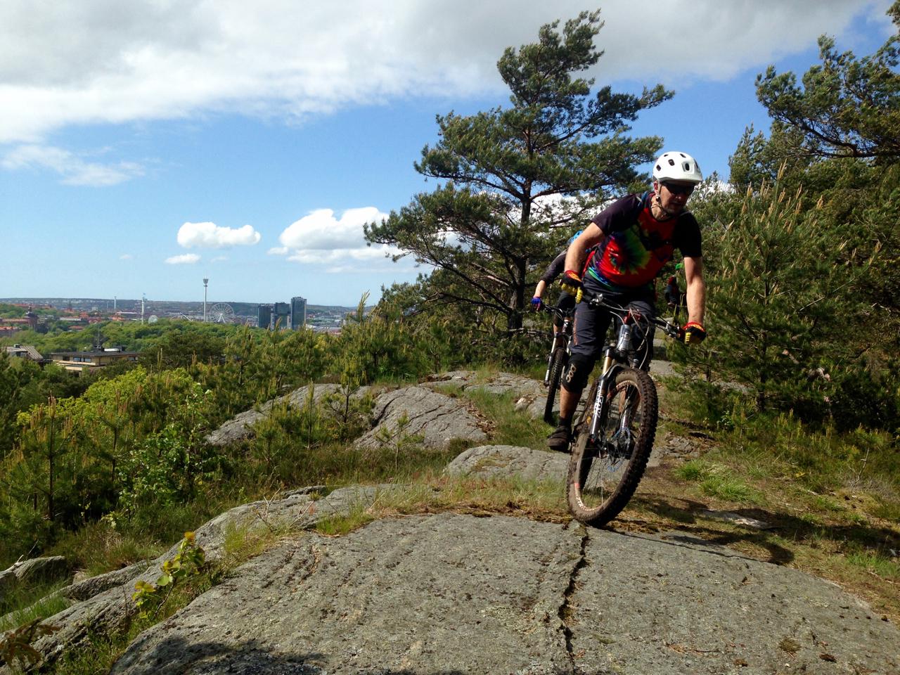 A mountain biker in a colorful shirt rides along a rocky trail, surrounded by lush greenery and trees. In the background, a city skyline can be seen under a partly cloudy sky. Molndal Area mountain bike trail.