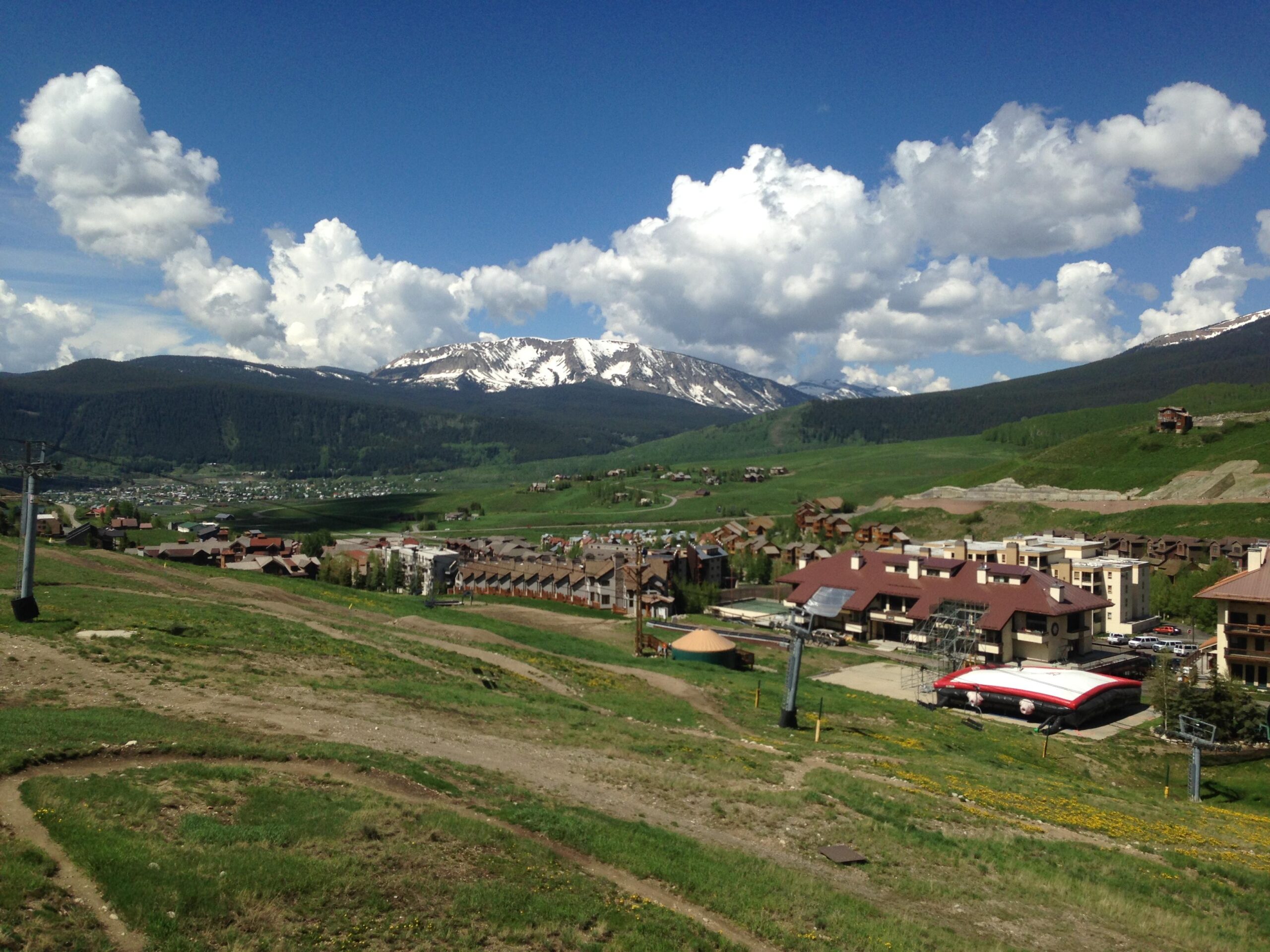 A scenic view of a mountainous landscape featuring lush green hills, a cluster of buildings at the base, and snow-capped peaks in the distance under a partly cloudy blue sky. A ski lift can be seen in the foreground, highlighting the area's recreational opportunities. Evolution Bike Park at Crested Butte Mountain Resort mountain bike trail.
