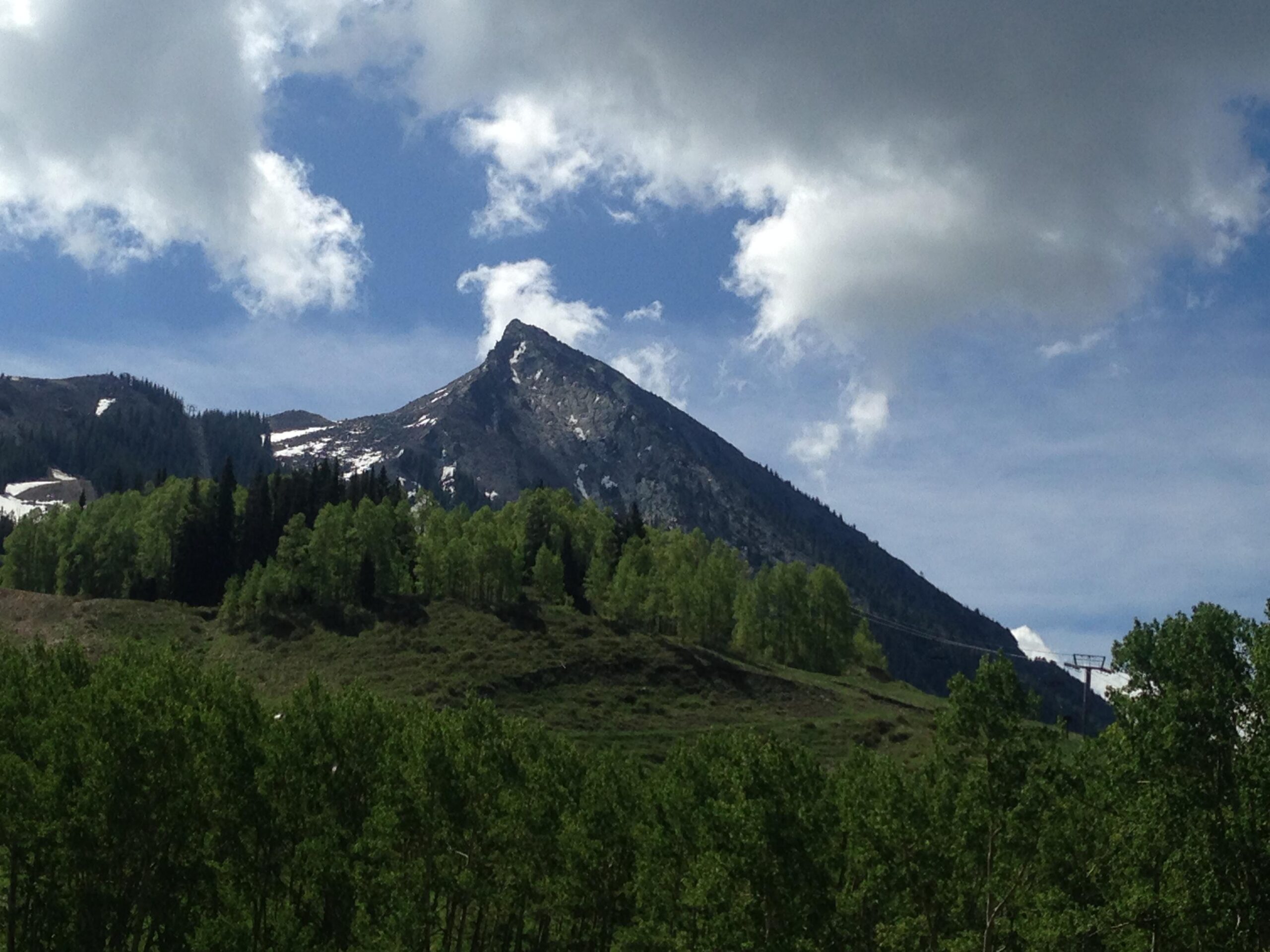 A scenic view of a mountainous landscape featuring a prominent peak against a backdrop of partly cloudy skies. In the foreground, lush green trees and hills are visible, while patches of snow can be seen on the mountain's surface. Evolution Bike Park at Crested Butte Mountain Resort mountain bike trail.