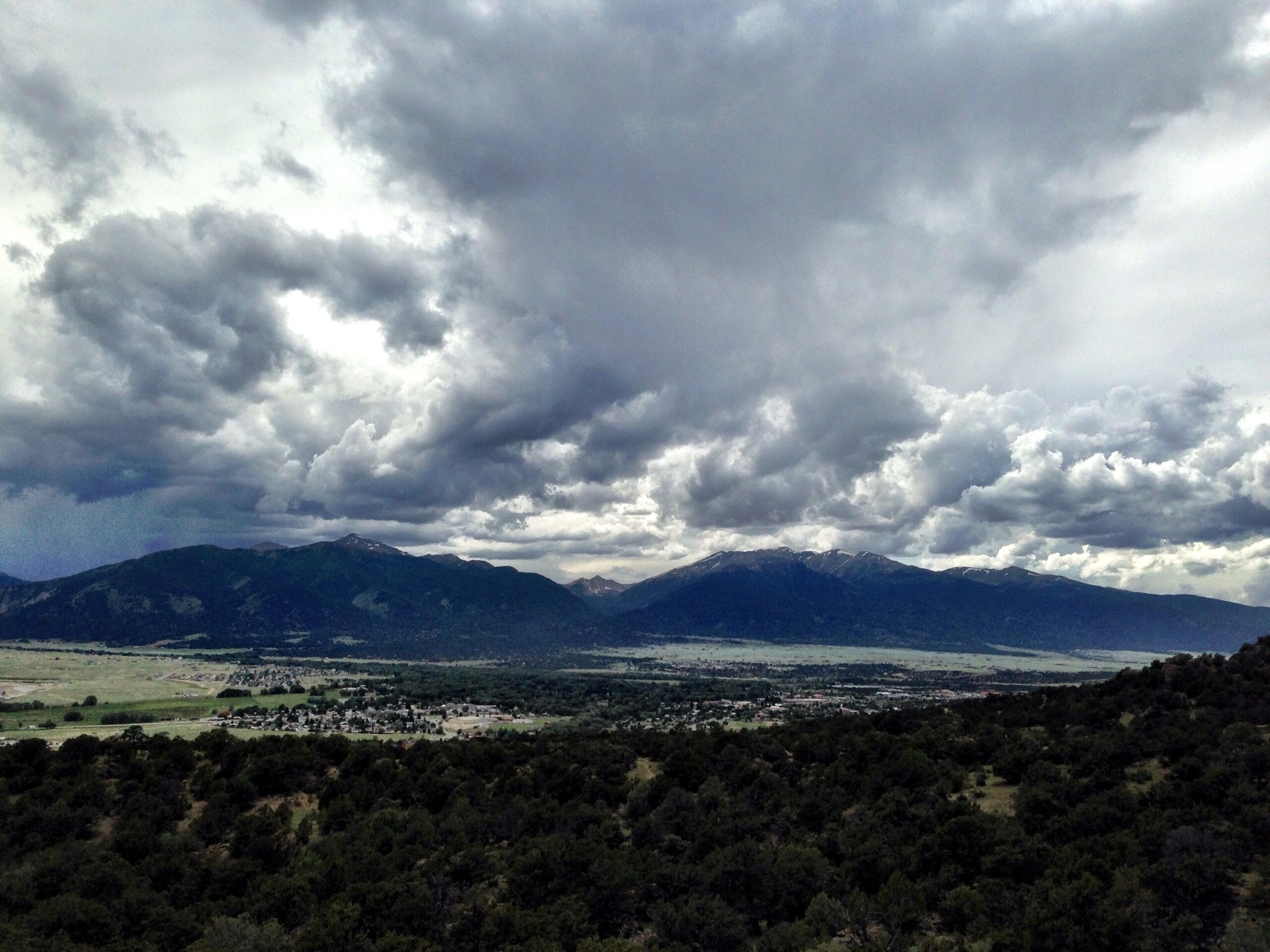 A panoramic view of a mountainous landscape under a dramatic sky filled with thick, swirling gray clouds. The foreground features a lush green valley and scattered trees, with a small town visible below. The mountains in the background are partially covered in snow, indicating elevation, while the clouds create a moody atmosphere over the scene. Midland Hills Trails mountain bike trail.