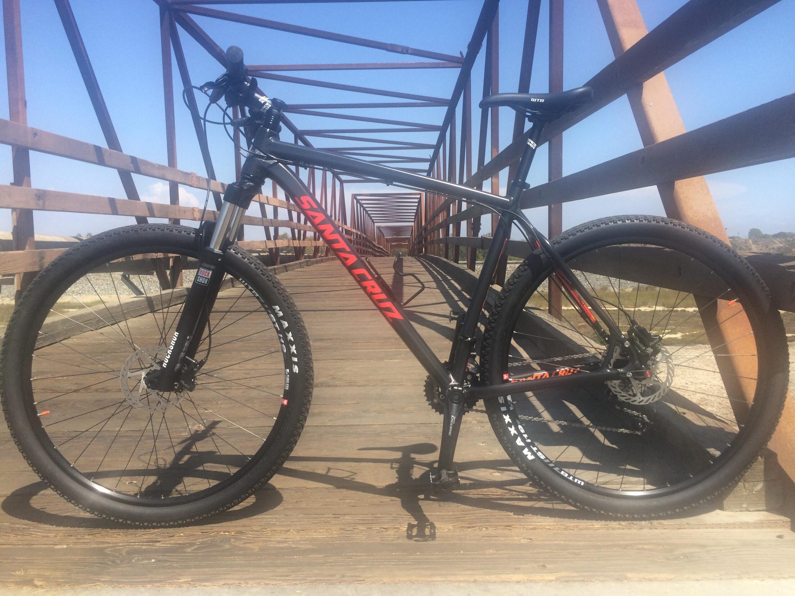 Santa Cruz Highball C: Mountain bike resting on a wooden bridge with a clear blue sky in the background. The bike features a black frame with red accents and is positioned at an angle to showcase its front wheel and suspension fork. The bridge's wooden structure and surrounding landscape are visible, adding context to the scene.