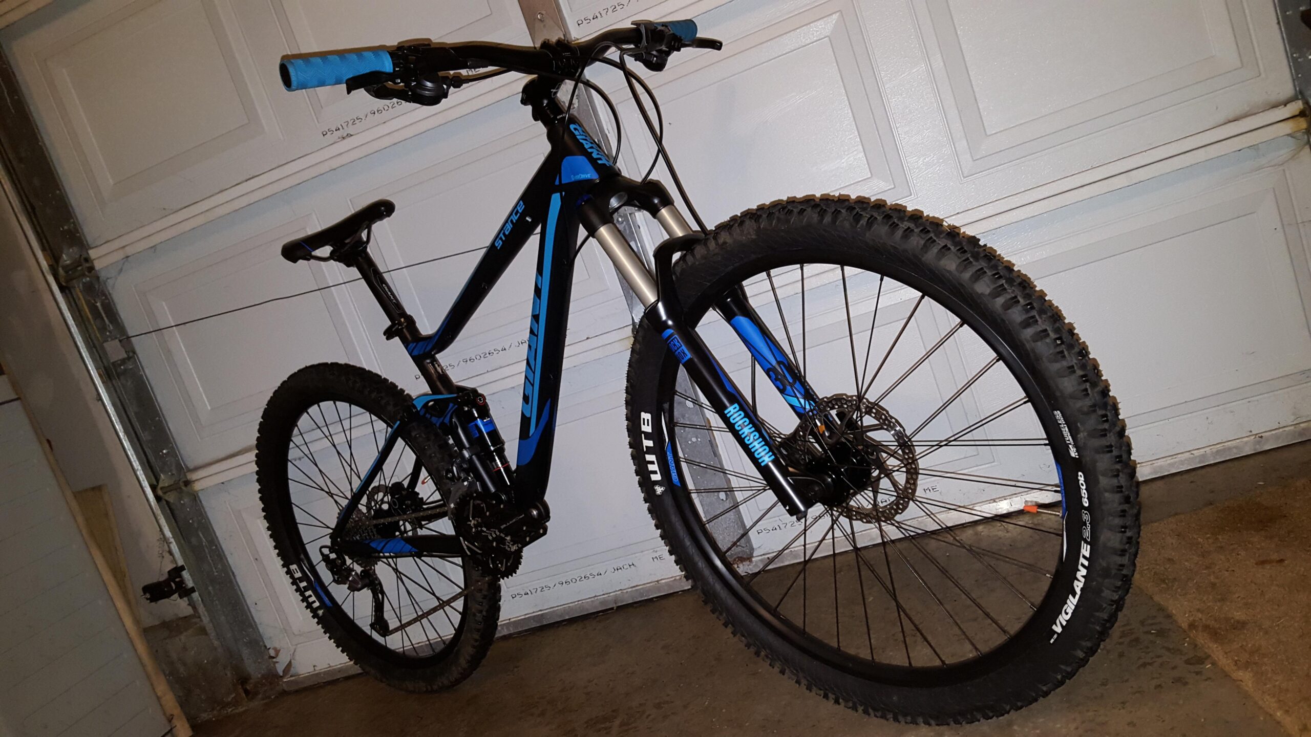 Giant Giant Stance 2: A black and blue mountain bike positioned against a garage door, featuring a sturdy frame, thick tires, and a front suspension fork. The bike displays blue handlebar grips and is set on a concrete floor.